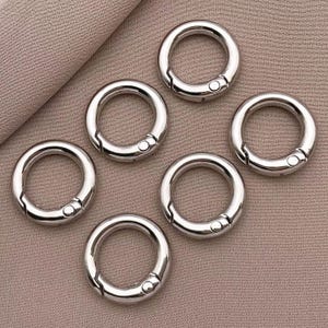 May include: Six silver metal key rings with a spring-loaded gate. The key rings are arranged in a circle on a light brown surface.
