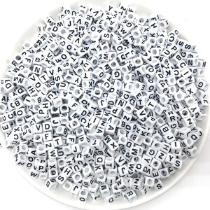May include: A white bowl filled with white plastic letter beads. The beads are all the same size and shape, and they have black letters on them. The letters are all different, and they are arranged randomly in the bowl.