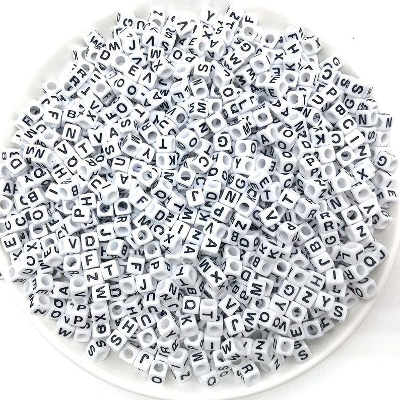 May include: A white bowl filled with white plastic letter beads. The beads are all the same size and shape, and they have black letters on them. The letters are all different, and they are arranged randomly in the bowl.