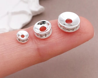 May include: Three silver-colored spacer beads of varying sizes. Each bead has a flat top and bottom with a central hole. The sides are adorned with small, sparkling embellishments, creating a decorative effect. The beads are displayed on a finger.