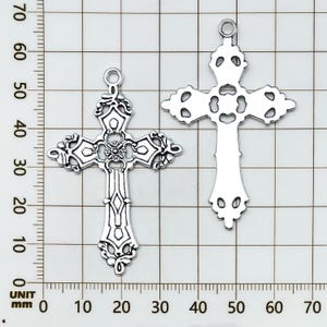 May include: Two silver-colored, ornate cross pendants. The crosses feature intricate filigree designs and a loop at the top for hanging. The crosses are displayed on a grid background, with measurements in millimeters.