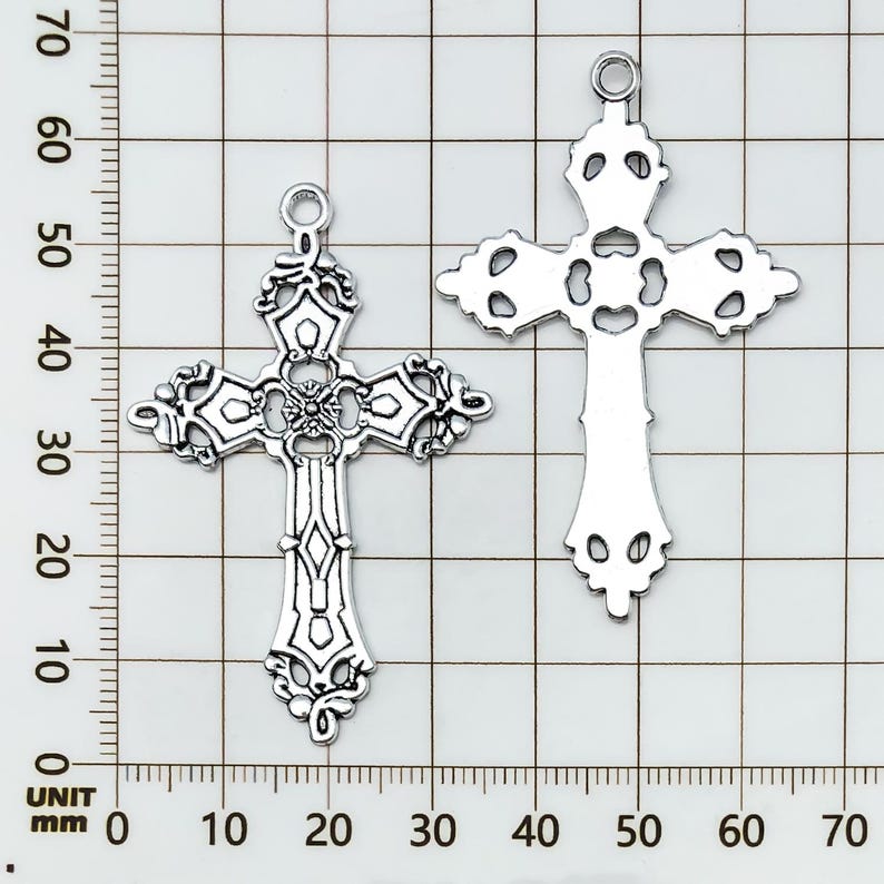 May include: Two silver-colored, ornate cross pendants. The crosses feature intricate filigree designs and a loop at the top for hanging. The crosses are displayed on a grid background, with measurements in millimeters.