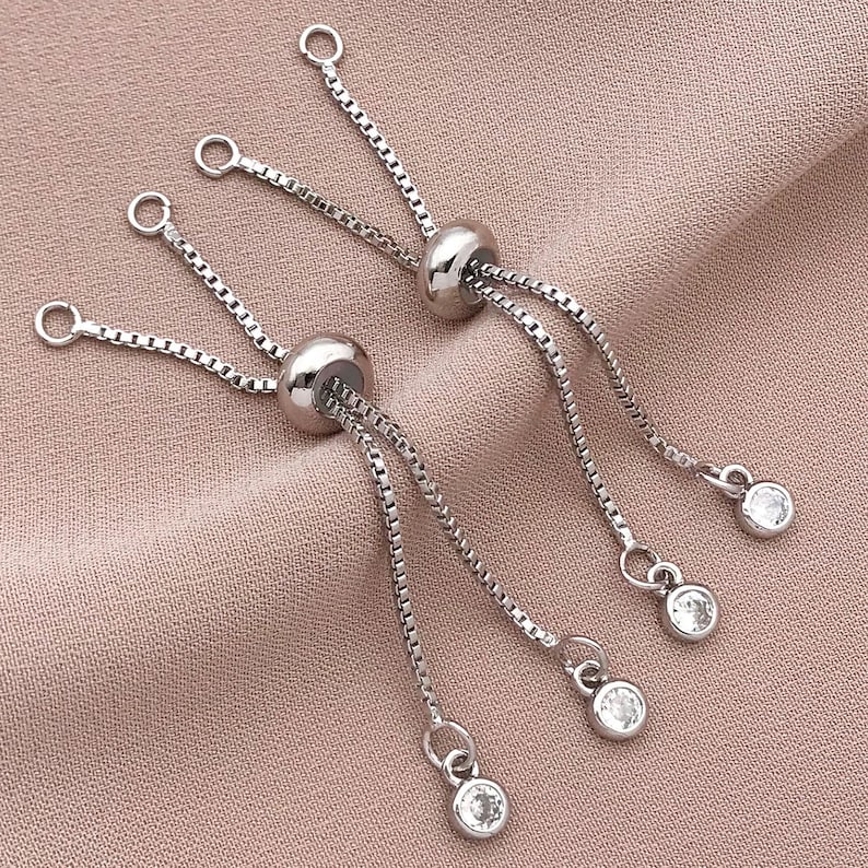 K&ouml;nnte beinhalten: Zwei verstellbare Silberarmb&auml;nder mit klaren Kristall-Akzenten. Jedes Armband hat eine verschiebbare Perle und mehrere klare Kristall-Anh&auml;nger.