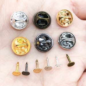 May include: A selection of metal pin backs in various colours, including silver, gold, and bronze. The pin backs are round with a butterfly clutch design. The image also shows the pin posts that attach to the backs.
