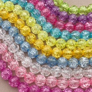 May include: Close-up of multiple strands of colorful, crackle-textured beads. The beads are arranged in rows, showcasing a vibrant spectrum of colors including pink, purple, blue, yellow, and green. The beads are round and have a textured, cracked appearance.