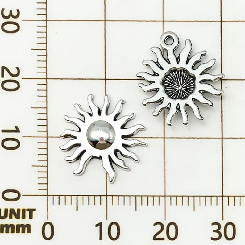 K&ouml;nnte beinhalten: Zwei silberfarbene Sonnenanh&auml;nger. Jeder Anh&auml;nger hat ein zentrales kreisf&ouml;rmiges Design mit strahlenf&ouml;rmigen Strahlen. Ein Anh&auml;nger hat eine Schlaufe zur Befestigung. Die Anh&auml;nger werden vor einem Rasterhintergrund mit Millimeterma&szlig;en angezeigt.