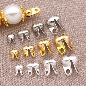 May include: Various small, tooth-shaped jewellery findings in gold and silver tones. Each finding has a loop at the base for attachment. A gold-toned clasp with a pearl bead is visible in the upper left corner. The background is a light pink fabric.