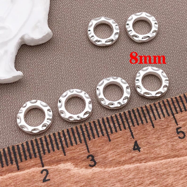 Pu&ograve; includere: Sei perline circolari argentate e strutturate, ciascuna di 8 mm di diametro, sono disposte su una superficie neutra. Un righello &egrave; posto vicino alle perline per la scala. Le perline hanno un design martellato.
