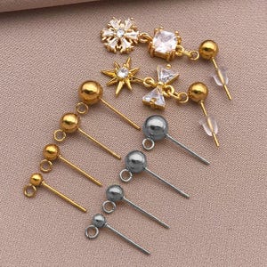 May include: An assortment of earrings in gold and silver tones. The earrings include stud styles with round and decorative elements, and dangle earrings with clear crystals and starburst designs. The earrings are displayed on a light pink surface.