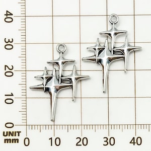 K&ouml;nnte beinhalten: Zwei silberfarbene Sternen-Charms. Jeder Charm hat eine Ansammlung von vier Sternen unterschiedlicher Gr&ouml;&szlig;e, die in der Mitte verbunden sind. Die Charms haben oben eine Schlaufe zum Aufh&auml;ngen. Der Hintergrund ist ein Raster mit Millimeter-Messungen.