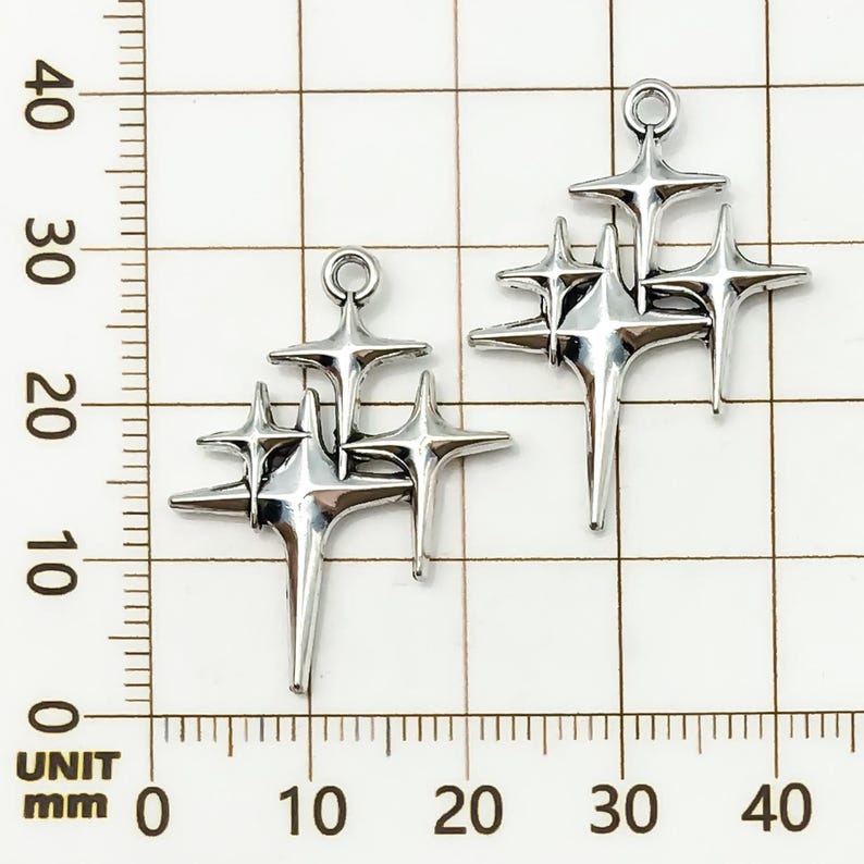 K&ouml;nnte beinhalten: Zwei silberfarbene Sternen-Charms. Jeder Charm hat eine Ansammlung von vier Sternen unterschiedlicher Gr&ouml;&szlig;e, die in der Mitte verbunden sind. Die Charms haben oben eine Schlaufe zum Aufh&auml;ngen. Der Hintergrund ist ein Raster mit Millimeter-Messungen.