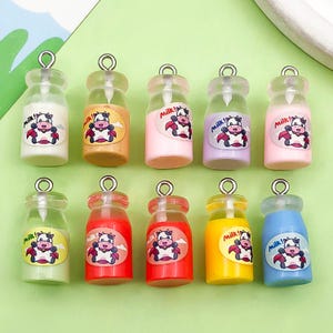 May include: A collection of eleven miniature milk bottle charms in various colors. Each bottle features a cartoon cow graphic and the text "Milk!" on a label. The bottles are clear with silver-colored loops at the top.