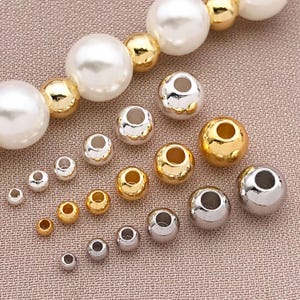 May include: A selection of beads in different sizes and finishes. The beads include large, off-white faux pearls, gold-coloured beads, and silver-coloured beads. The beads are arranged on a textured, light brown surface, displaying their different sizes and metallic sheens.