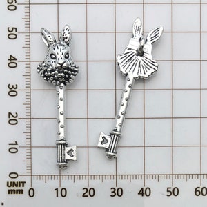 May include: Two silver-toned key charms, one with a rabbit head and the other with a stylized rabbit head wearing a crown. Both charms have a keyhole and a decorative key stem.