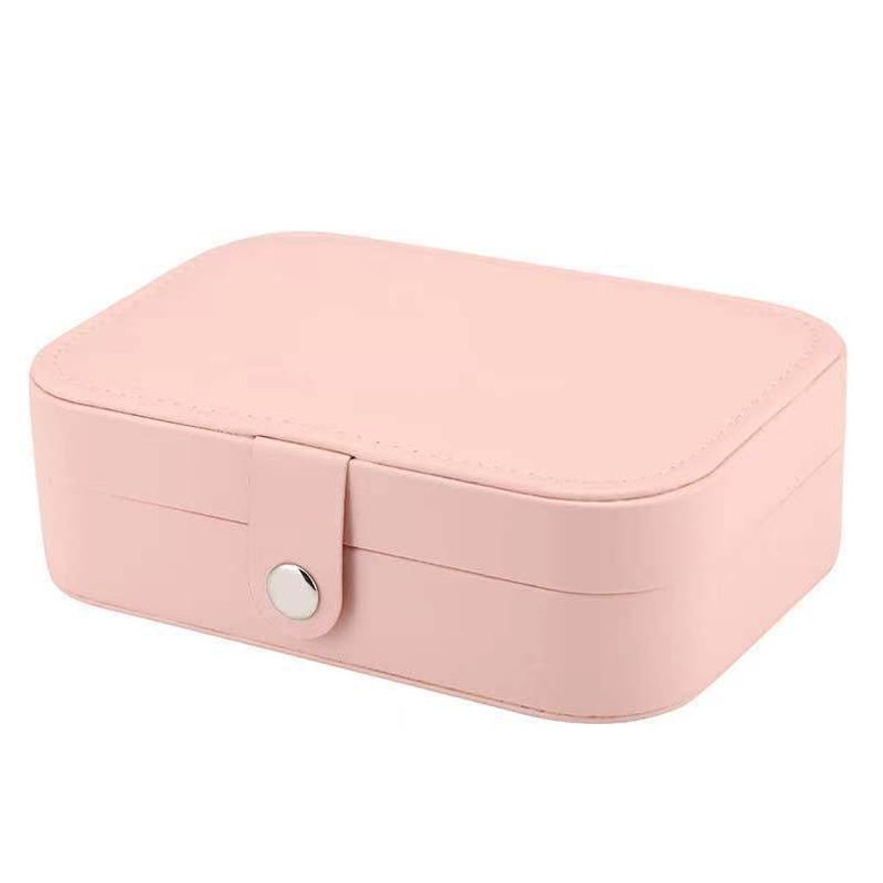 May include: A pink faux leather jewellery box with a silver button closure. The box has two compartments for storing jewellery.