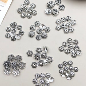 May include: Assortment of silver-toned metal bead caps with intricate floral designs. The caps are arranged in groups on a white surface.