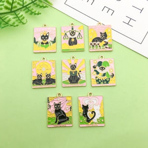 May include: A selection of rectangular tarot card charms in pink and gold, each with a black cat design. The charms represent various tarot cards, such as The Justice, The Lovers, and The Fool. Each charm has a gold-coloured loop at the top for attachment.
