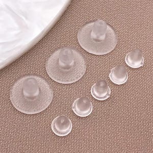 Puede incluir: Un conjunto de botones de silicona transparentes. Los botones, de diferentes tamaños, tienen una base circular plana y un centro redondeado en relieve. Están dispuestos sobre una superficie texturizada de color beige, sugiriendo un contexto artesanal o de costura.