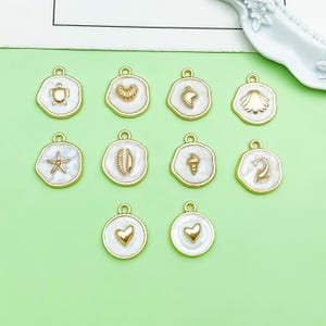 May include: A collection of ten circular charms featuring white enamel centers and gold-toned metal rims. Each charm displays a unique raised gold-toned icon, including a turtle, various seashells, a starfish, a dolphin, and two heart shapes.