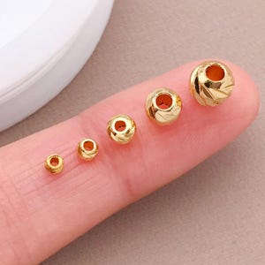 May include: Five small, gold-coloured beads of varying sizes. Each bead has a hole through the centre and a textured surface. The beads are displayed on a finger, showcasing their size and detail.