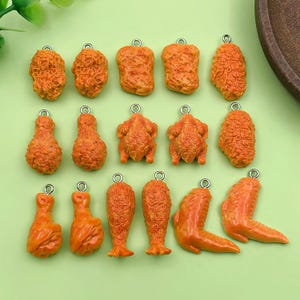 Peut inclure: Une collection de breloques en forme de poulet frit orange, comprenant des pilons, des ailes et des morceaux de poulet. Chaque breloque a une petite boucle argentée en haut pour l'attacher à des bijoux ou autres créations. Les breloques sont disposées sur une surface vert clair.