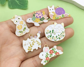 May include: A set of seven gold-tone metal charms featuring various animal and nature designs. Each charm is white with colorful enamel details, including a cat, a turtle, a snail, a fox, a rabbit, and two additional cat charms.