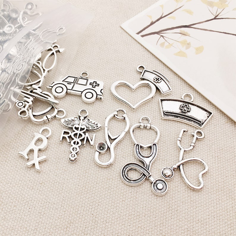 May include: A collection of silver-toned metal charms with medical themes, including a stethoscope, a heart, a cross, a pill bottle, an ambulance, and a nurse's cap.