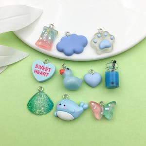 Peut inclure: Assortiment de 10 breloques en résine colorées de formes et de motifs variés, dont un nuage, une empreinte de patte, un cœur, une coquille, une baleine, un papillon, une barre chocolatée, une bouteille de thé à bulles et un canard. Les breloques sont toutes de couleur bleu clair, rose ou verte.