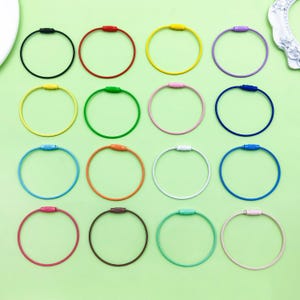 May include: A collection of colorful keychains in various shades, including black, red, yellow, purple, green, pink, blue, orange, white, and brown. Each keychain features a circular design with a matching colored clasp, arranged on a light green surface.