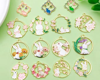 May include: A collection of gold-toned metal charms featuring white rabbits and floral designs. The charms are in various shapes, including circles and diamonds, with pink, green, and blue accents. These decorative charms are ideal for jewelry making or crafting.
