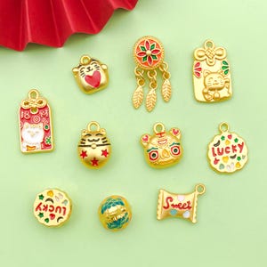 May include: Assortment of gold-colored charms with enamel details. Charms feature various designs including a cat, a lion, a dream catcher, a candy bar, a bell, and a lucky charm.