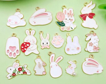 May include: A collection of white and gold-toned enamel bunny charms in various poses. Some charms feature pink accents, flowers, or bows. The charms are designed with a gold outline and a loop for attaching to jewelry or crafts.