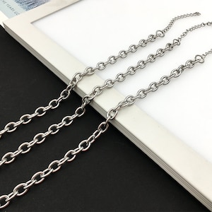 Puede incluir: Tres collares de cadena plateados con eslabones ovalados se muestran sobre un fondo blanco y negro. Las cadenas están dispuestas en diagonal, mostrando su diseño y textura. Las cadenas están hechas de un metal brillante.