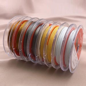 May include: A collection of spools of colorful beading wire in a clear plastic holder. The wires are in various colors, including gold, silver, yellow, and white. The spools are arranged in a semi-circle, with the ends of the wire visible.