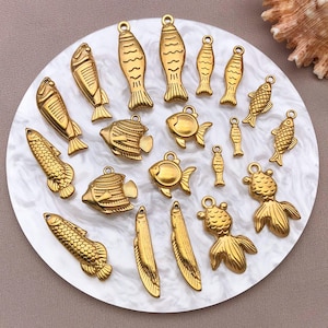 May include: A collection of gold-colored fish charms in various designs, including detailed scales and fins. The charms are arranged on a white and gray marbled surface, showcasing their intricate details and shapes. These charms are ideal for jewelry making.