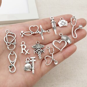May include: A collection of silver-toned metal charms featuring medical symbols, including a stethoscope, a heart, a syringe, a vial, an Rx symbol, and a caduceus. The charms are perfect for jewelry making or crafting.