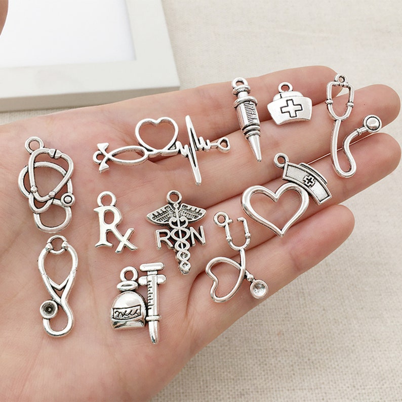 May include: A collection of silver-toned metal charms featuring medical symbols, including a stethoscope, a heart, a syringe, a vial, an Rx symbol, and a caduceus. The charms are perfect for jewelry making or crafting.