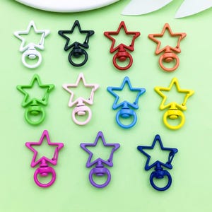 Puede incluir: Una colección de cierres giratorios en forma de estrella en varios colores: blanco, negro, rojo, naranja, verde, rosa, azul, amarillo, morado y azul marino. Cada cierre tiene un anillo para sujetar.