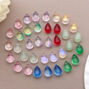 May include: A collection of teardrop-shaped beads in various colors, including pink, blue, green, and yellow. The beads are transparent and appear to be made of glass or a similar material. They are arranged on a neutral-toned surface.