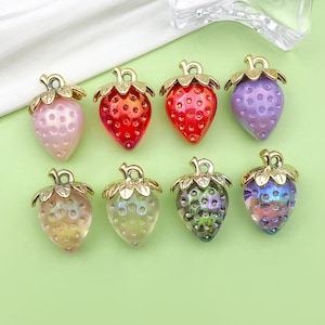 May include: Eight strawberry-shaped charms in various colors, including pink, red, purple, yellow, and iridescent. Each charm has a gold-colored stem and leaves. These charms are likely for jewelry making or crafting.