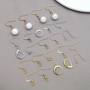Può includere: Un assortimento di ganci e componenti per orecchini color oro e argento. Alcuni ganci hanno un design curvo semplice, mentre altri presentano una piccola sfera o una chiusura rotonda. Sono presenti anche diversi orecchini con perline bianche e rotonde.