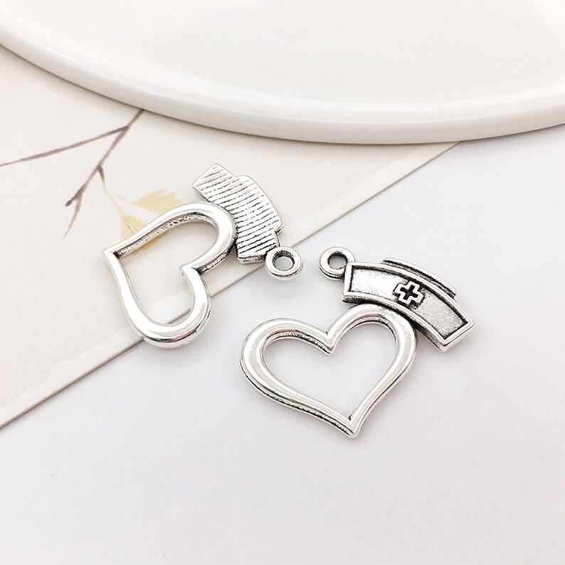 May include: Two silver charm pendants, each featuring a heart-shaped frame with a nurse's cap on top. The caps have a cross design.