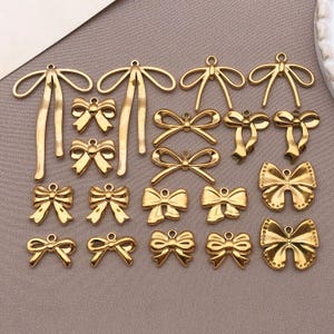 Könnte beinhalten: Eine Sammlung goldfarbener Schleifenanhänger in verschiedenen Designs. Einige Schleifen haben lange, fließende Bänder, während andere einfach und kompakt sind. Die Anhänger sind auf einer neutral getönten Oberfläche angeordnet, die ihre komplizierten Details und ihr poliertes Finish hervorhebt.