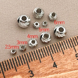 May include: A selection of silver beads in various sizes, from 2.5mm to 6mm. Some beads feature a spiral design with white detailing. A wooden ruler is in the foreground.
