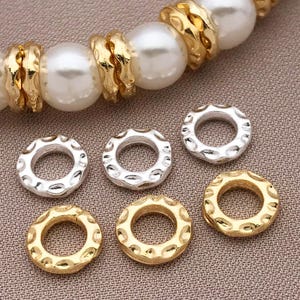 Pu&ograve; includere: Primo piano di componenti di gioielli, tra cui perline ad anello strutturate in oro e argento e una collana di perle. Le perline ad anello hanno un design strutturato e ondulato. Le perle sono grandi e bianche.