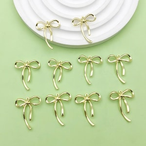 Könnte beinhalten: Zehn goldfarbene Metall-Schleifen-Anhänger. Die Schleifen sind alle gleich groß und gleich geformt und haben ein leicht verdrehtes Design. Die Anhänger sind perfekt, um Schmuck, Bastelarbeiten und anderen Projekten einen Hauch von Eleganz zu verleihen.