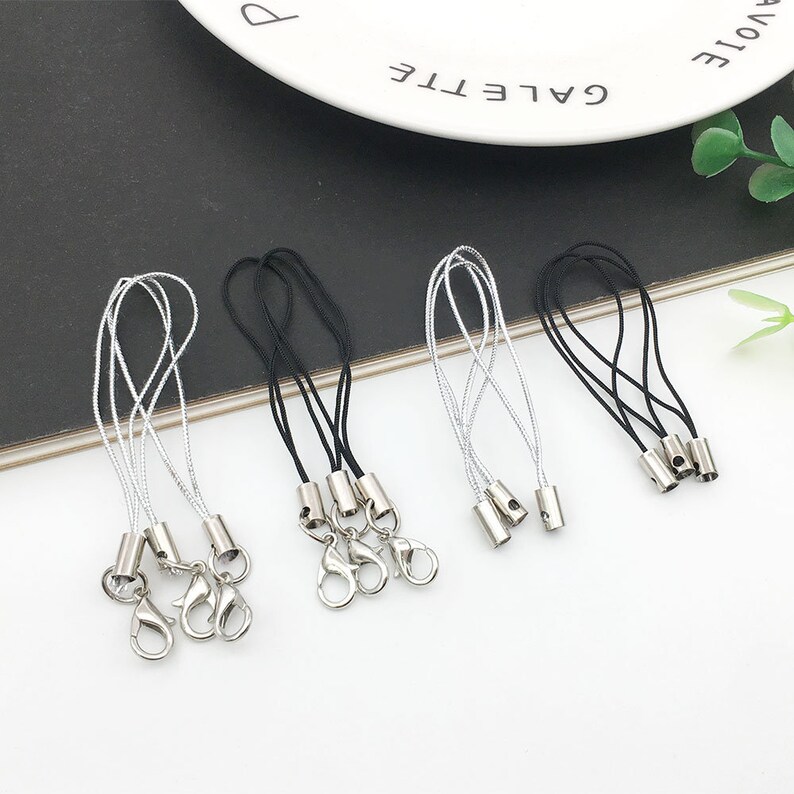 Op de afbeelding: Zes zilveren sleutelhangers met zwarte en zilveren koorden. De sleutelhangers hebben een zilveren lus en een zilveren tonvormige sluiting.