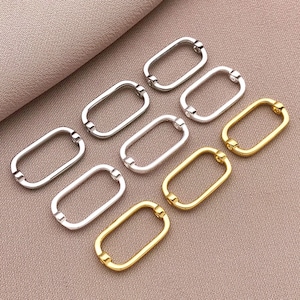 May include: A collection of oval-shaped metal clasps in silver and gold tones. The clasps are arranged on a neutral-colored surface, showcasing their design and finish. These clasps are likely used for jewelry making or crafting projects.