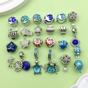 May include: A collection of 20 silver charm beads featuring a variety of sea life designs, including starfish, seashells, seahorses, crabs, turtles, and fish. The beads are decorated with colourful enamel and some have sparkling rhinestones.