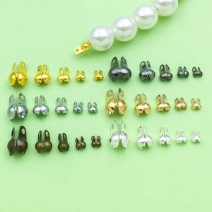 May include: A selection of metal bead crimp end findings in various sizes and colours, including gold, silver, and bronze. The findings are arranged on a light green surface, with a strand of white pearls in the background.